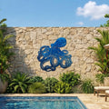 Blue octopus sculpture on a stone wall by a pool with plants and a clear sky.