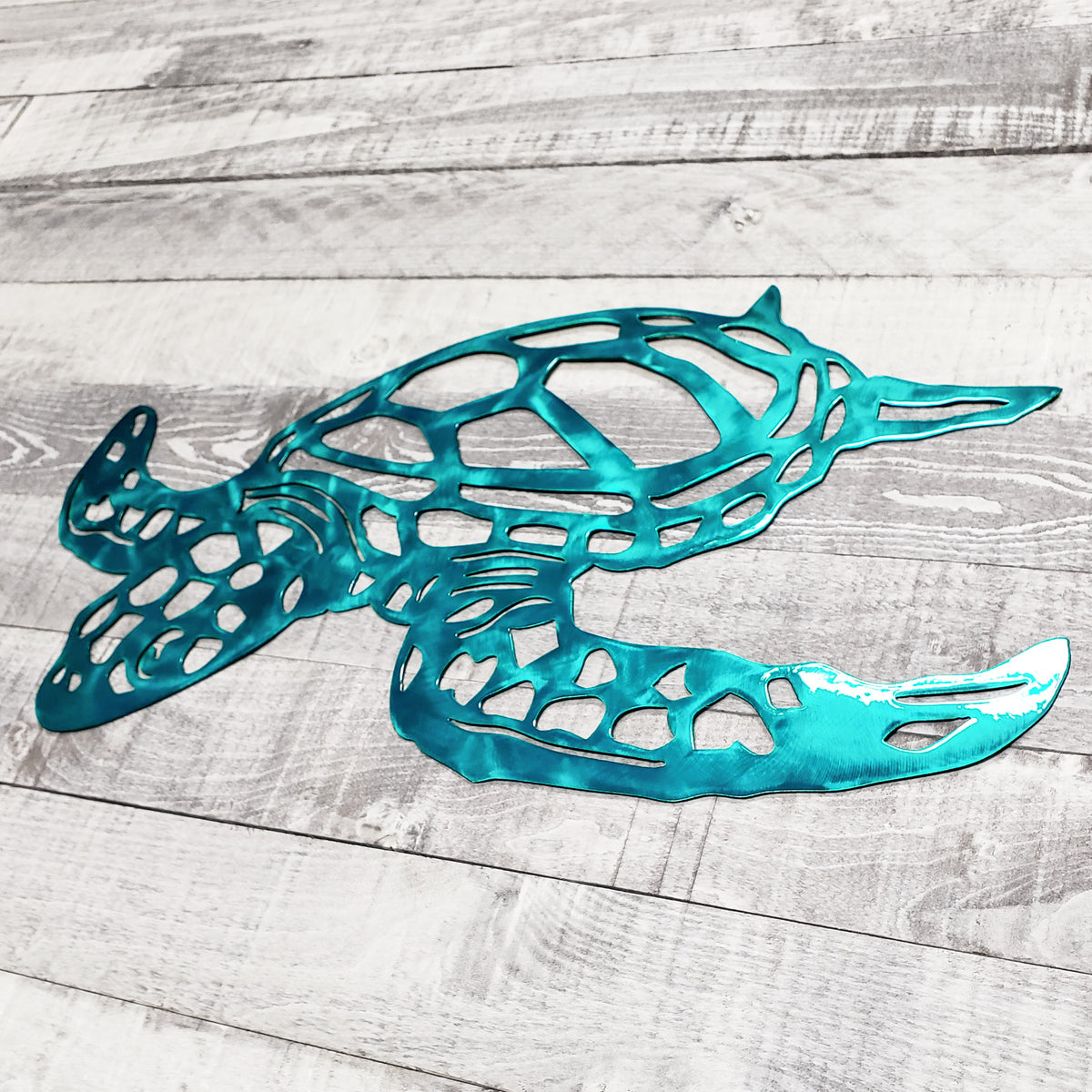A teal metallic cutout wall art piece shaped like a sea turtle, displayed on a light gray wood plank surface.