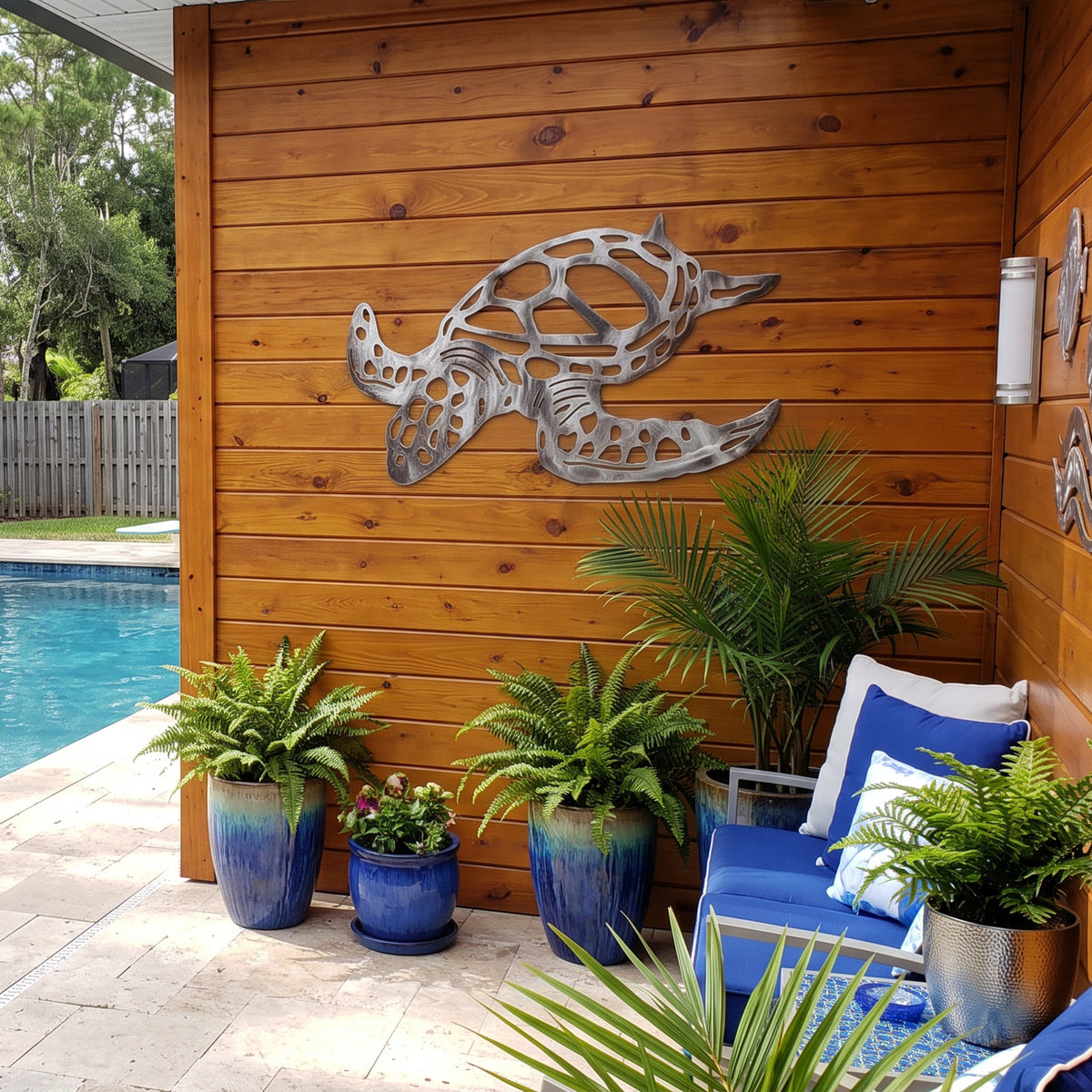 Metal turtle wall art mounted on a warm wood-paneled wall in a backyard patio setting with blue lounge chairs, potted ferns, and a swimming pool visible.
