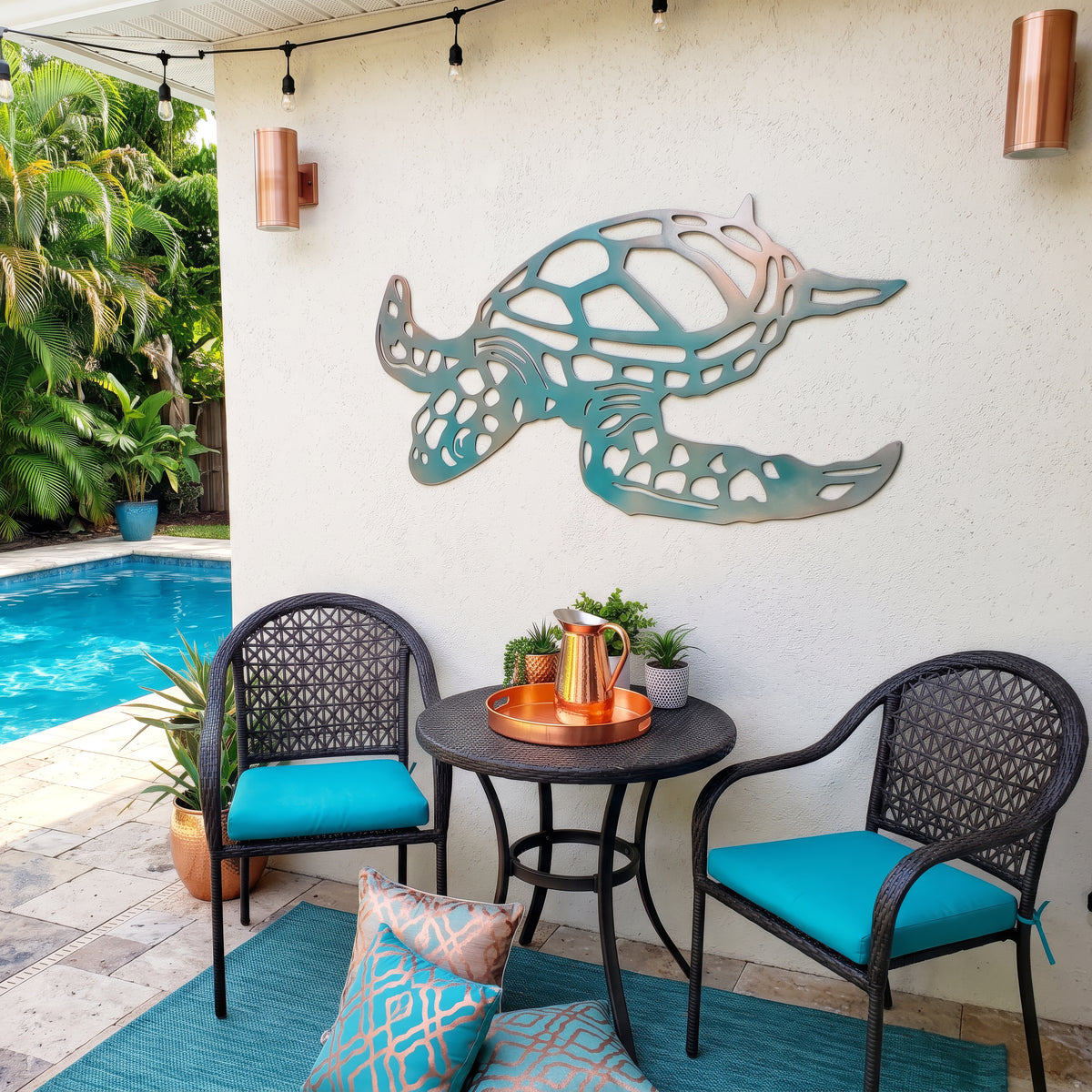 A teal and light copper metal turtle wall art piece mounted on a white stucco wall in a backyard patio setting. The patio features two black wicker chairs with bright turquoise cushions and a small round black table with a copper tray.